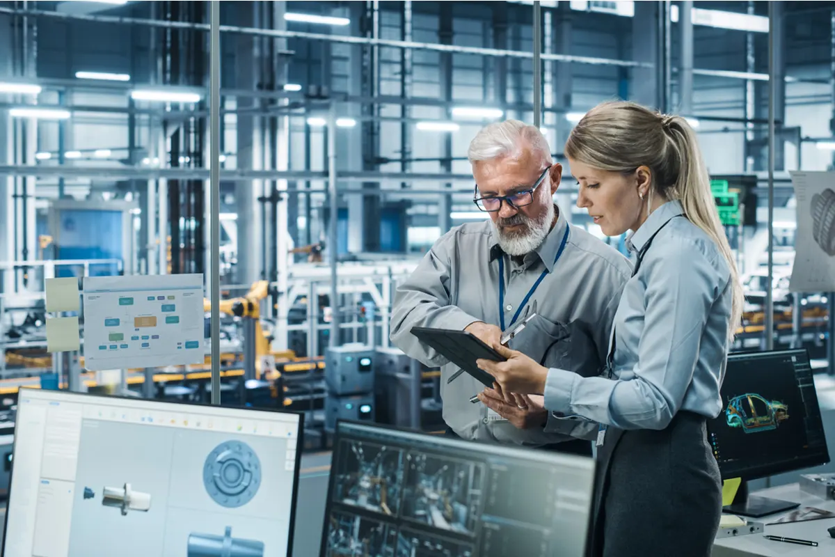 Two engineers in a manufacturing plant reviewing CAD drawings on a tablet connected to a high-speed network